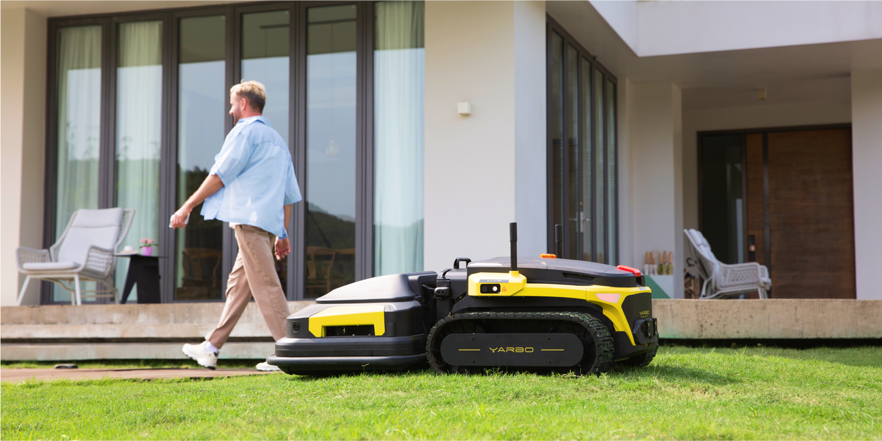 Yellow and black robotic lawn mower on green grass near modern home, man walking by casually.