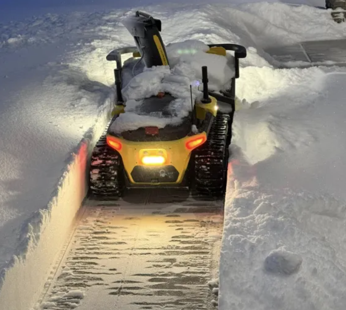 Yellow Yarbo snow blower clears deep snow at night, headlights glowing, tracks carving a clean path through winter.