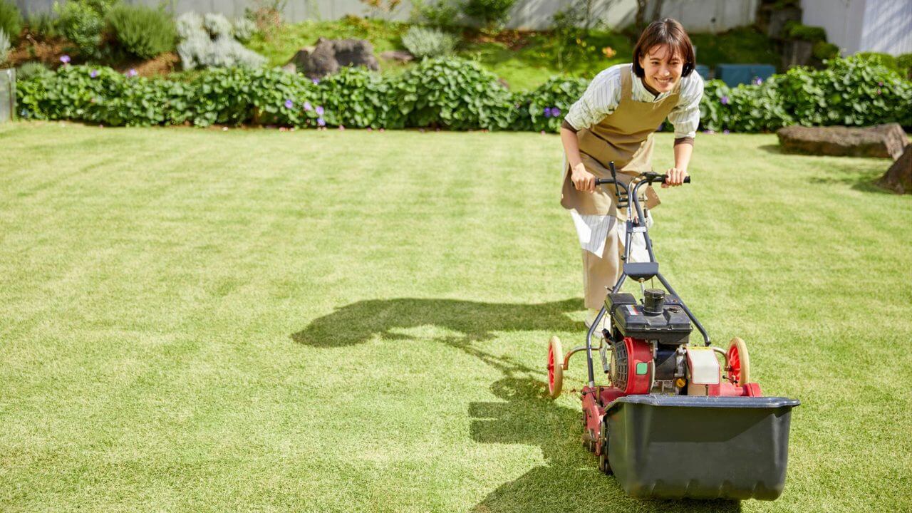 Woman smiling while pushing a red lawnmower across a lush, evenly cut green lawn on a sunny day.