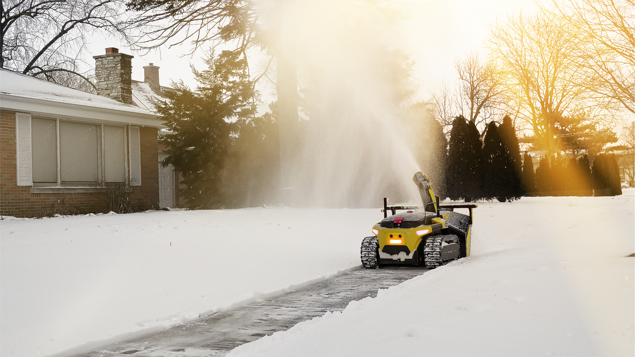 Yellow two-stage snow blower clears driveway in snowy yard at golden hour, powerful snow plume rising.