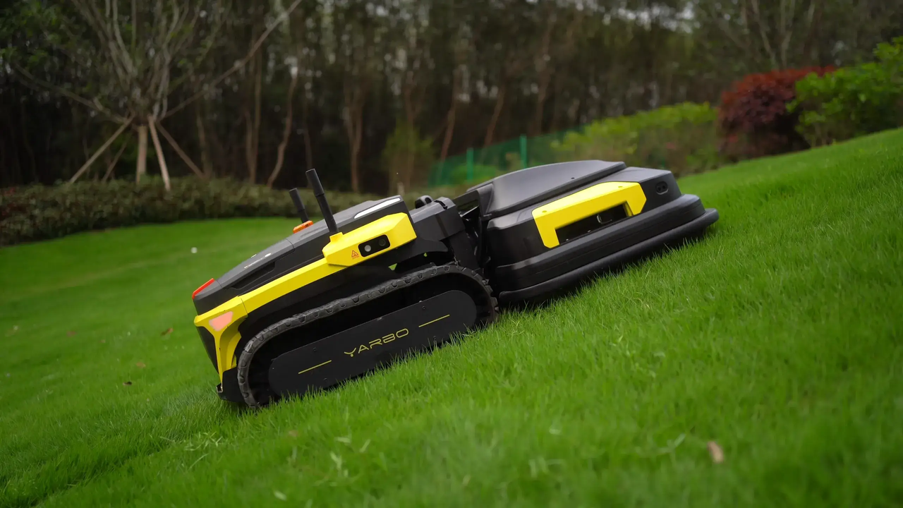 Yellow and black robotic lawn mower with tank treads navigating a grassy slope in a lush garden.