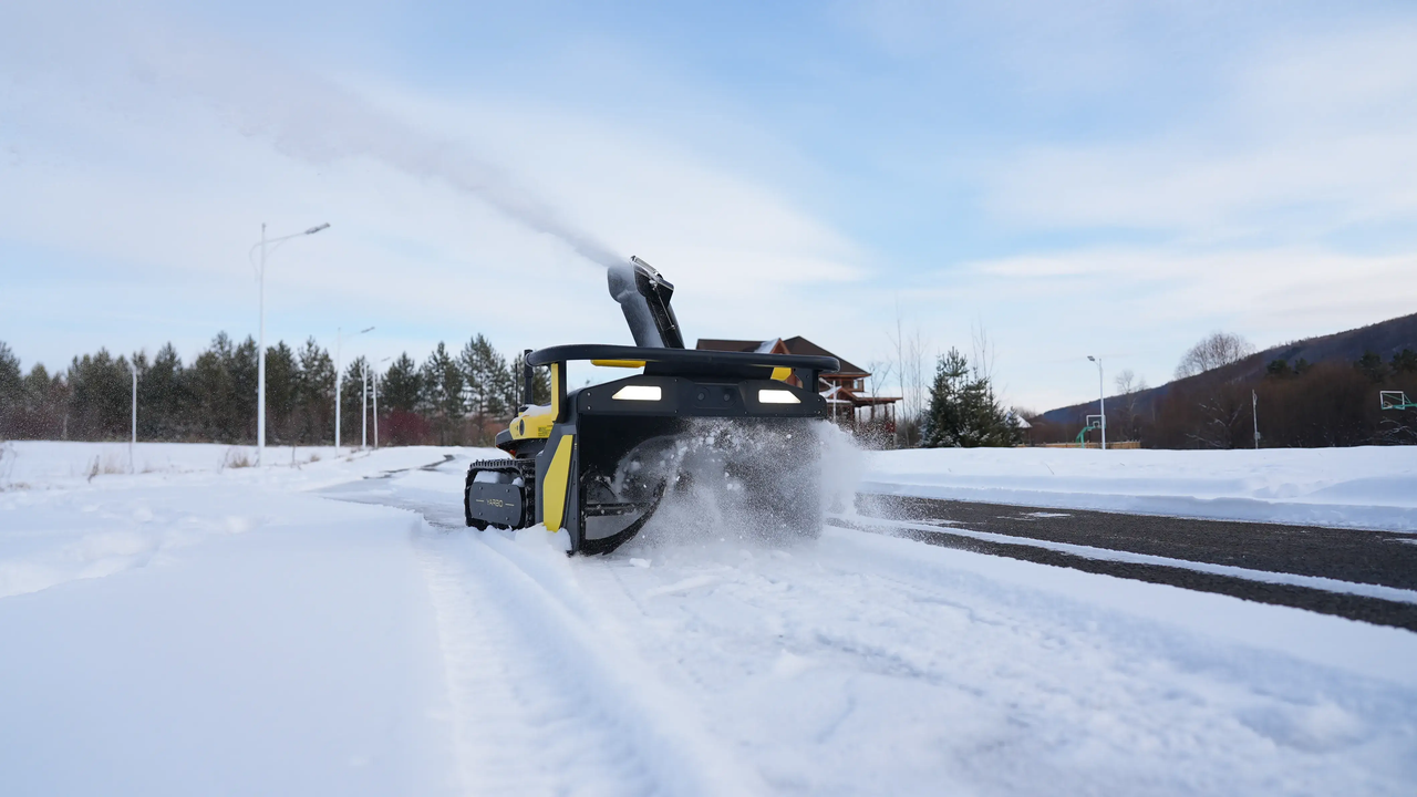 Yellow and black snow blower clearing a snow-covered driveway on a bright winter day, with trees and a house in the background.