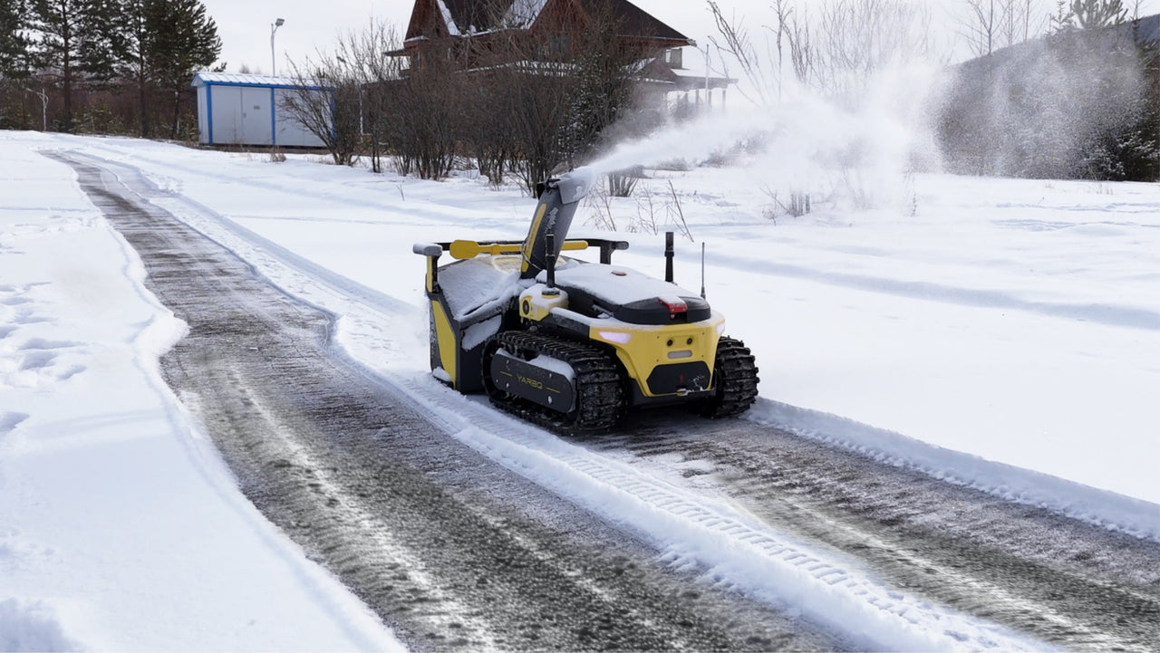 Yellow Yarbo snow blower clears compacted snow from a driveway, revealing clean pavement beneath.