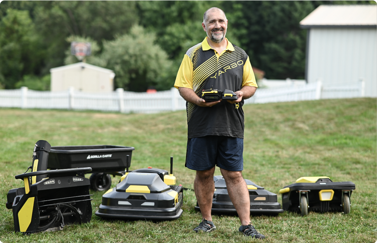 Smiling man in Yarbo shirt stands on lawn with robotic mowers, holding remote, showcasing smart lawn care technology.