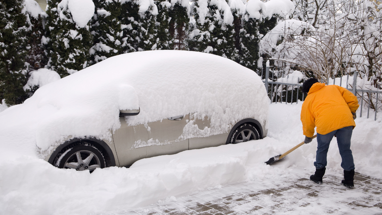 Person in bright yellow coat shoveling snow from driveway beside snow-covered car, winter scene.