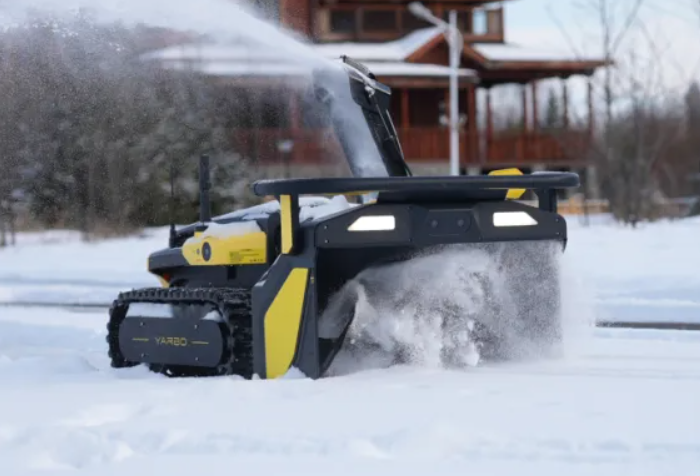 Yellow and black robotic snowblower clearing snow in winter yard, spraying snow sideways near wooden house.