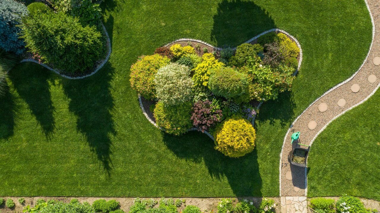 Aerial view of a lush, manicured lawn with colorful garden beds, winding stone path, and a green lawn mower—symbolizing precision and care in lawn maintenance.