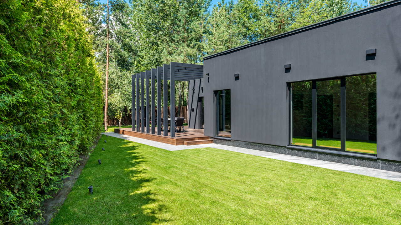 Lush green lawn beside modern gray house with wooden deck and pergola, showcasing post-mow lawn care results.