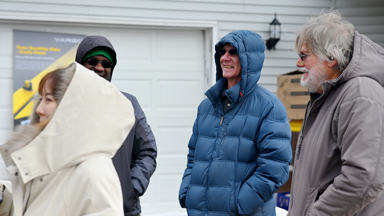 Four people in winter coats laugh together outside a house, with a Yarbo tour poster visible in the background.