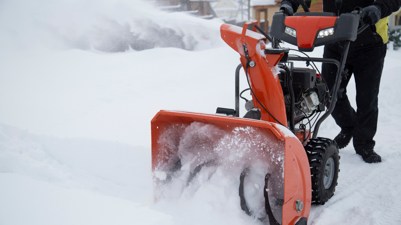 Person operates bright orange self-propelled snow blower, clearing deep snow from driveway on cold winter day.