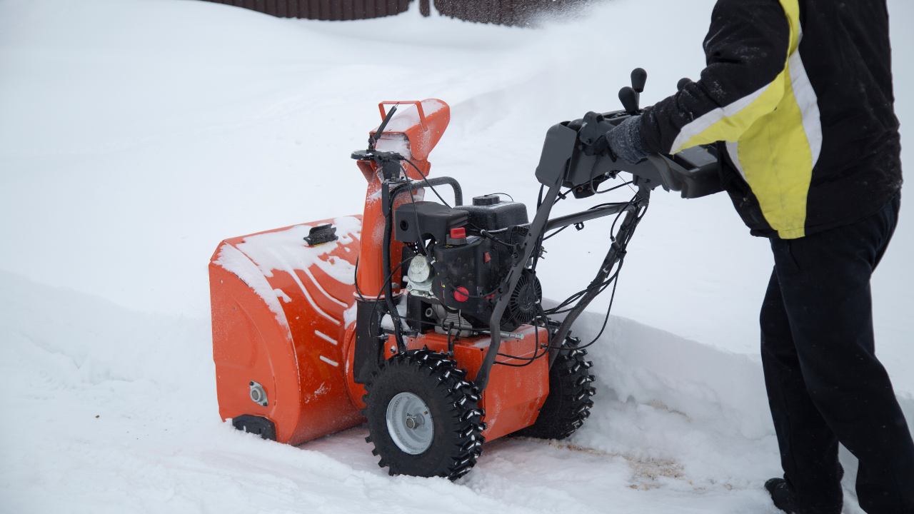 Person operating orange gas-powered snow blower in deep snow, clearing driveway with visible engine and rugged tires.