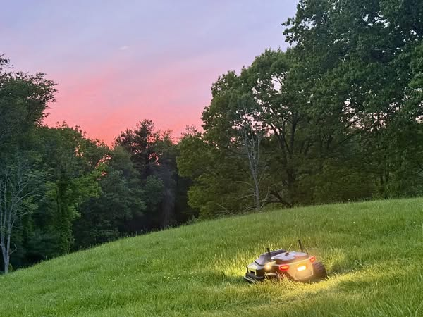 Robotic lawn mower glows on lush green hill at sunset, symbolizing smart, efficient yard care.