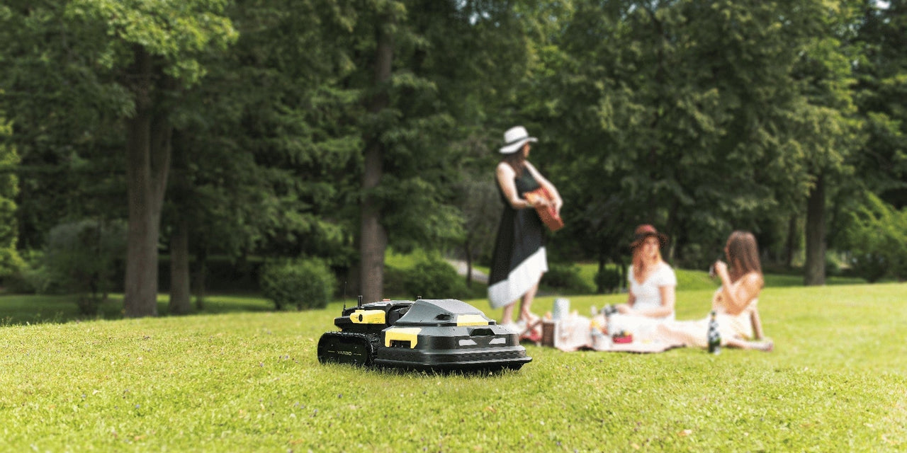 Robot lawn mower on vibrant green grass, with women enjoying a picnic in the background under leafy trees.