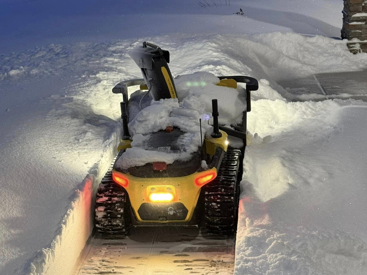 Yellow snow blower clearing deep snow at night, headlights glowing, tracks cutting through winter landscape.