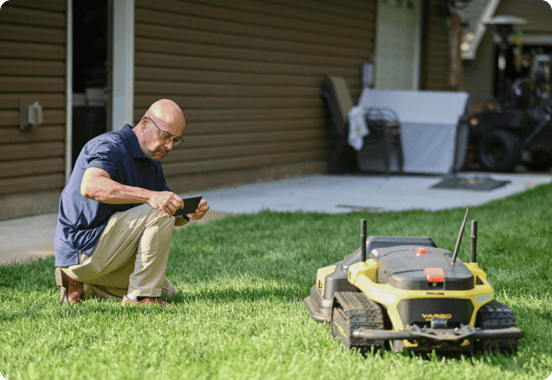 Man kneels on lawn, checking smartphone while yellow robotic mower operates nearby, showcasing smart lawn care technology.