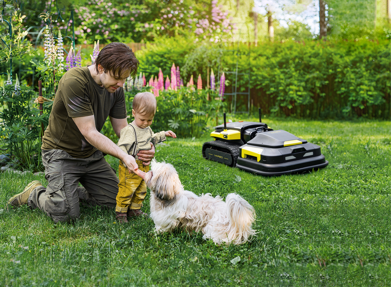 Father and child play with dog in lush garden while robotic lawn mowers work quietly in background.