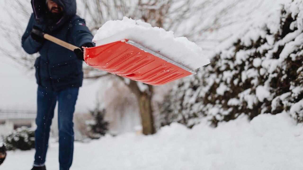 Man in winter coat shovels snow with red shovel in snowy yard, emphasizing safe snow removal.