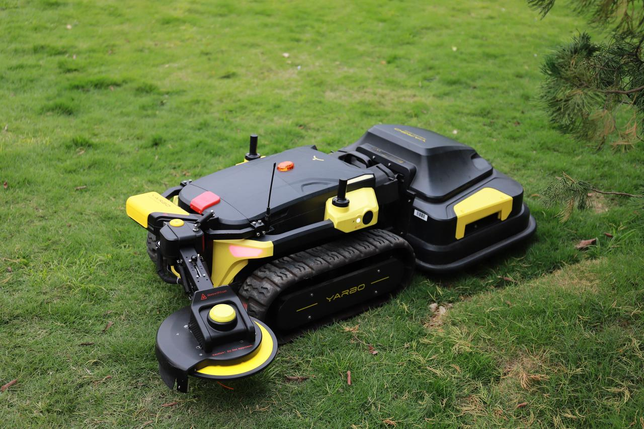 Black and yellow robotic lawn mower with tank treads and trimmer attachment, resting on green grass near pine branches.