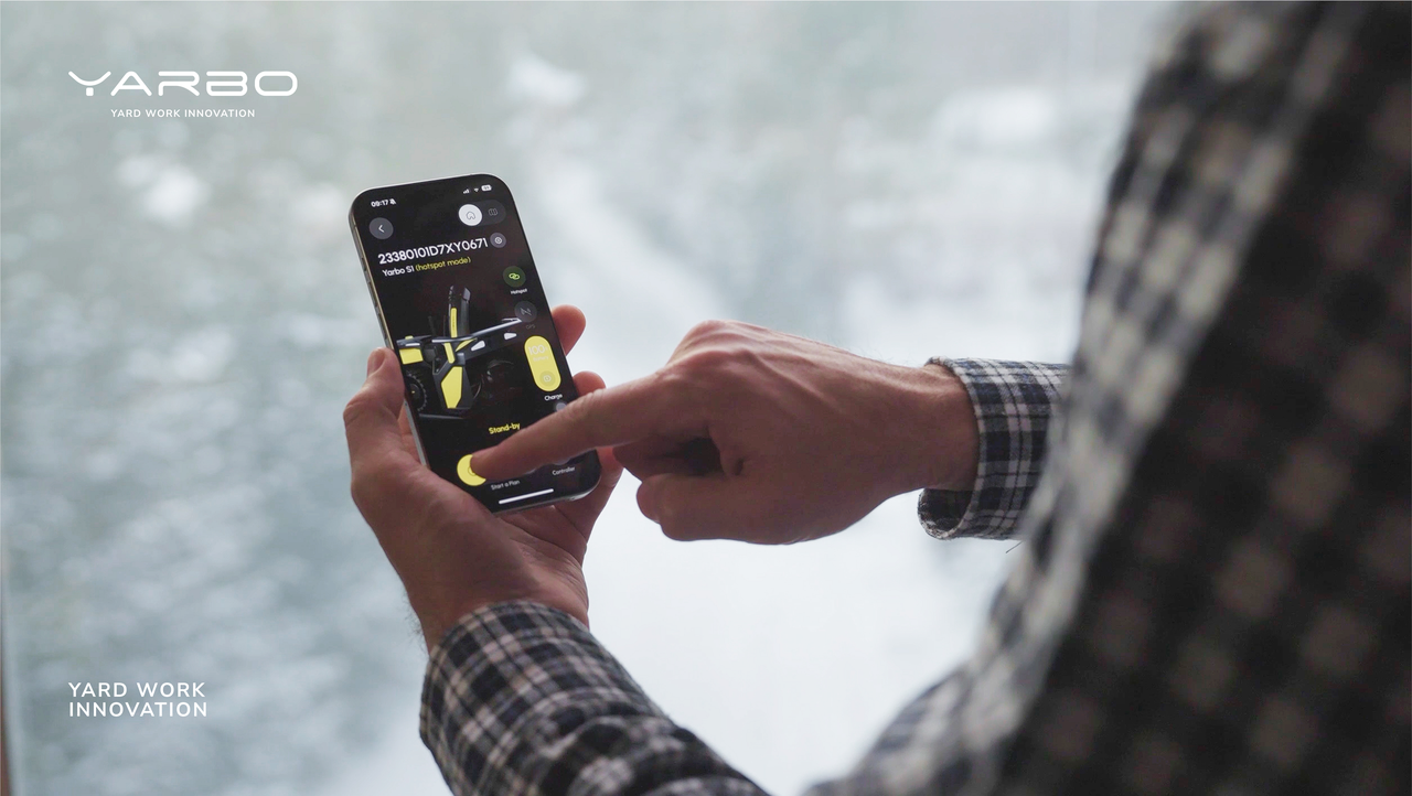 Hands operate Yarbo snowblower via smartphone app, snow-covered yard in background.