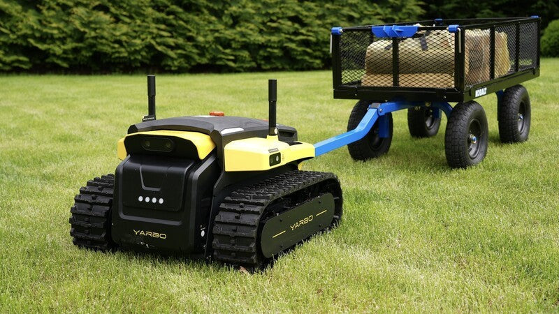 Black and yellow Yarbo robot mowing lawn, towing blue utility wagon with cargo, showcasing attachment compatibility.
