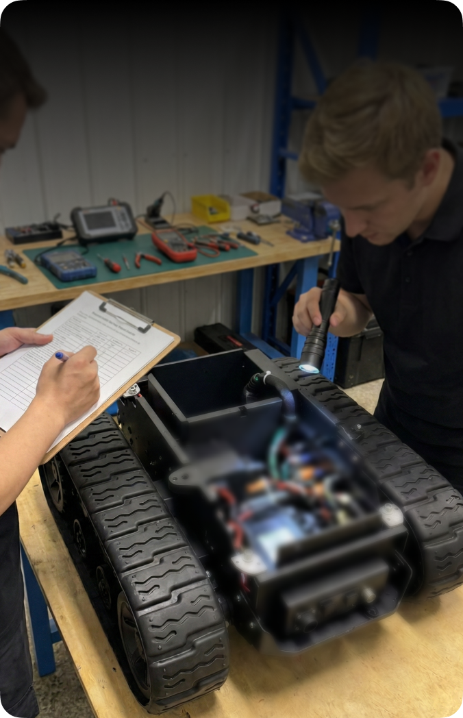 Two engineers inspect a tracked robot on a workbench; one uses a flashlight, the other writes notes on a clipboard. Tools and electronics visible in background.