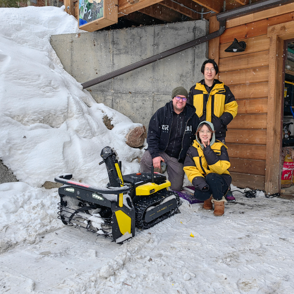 Three people in yellow and black jackets pose with a yellow and black tracked robot in snowy yard beside wooden cabin.
