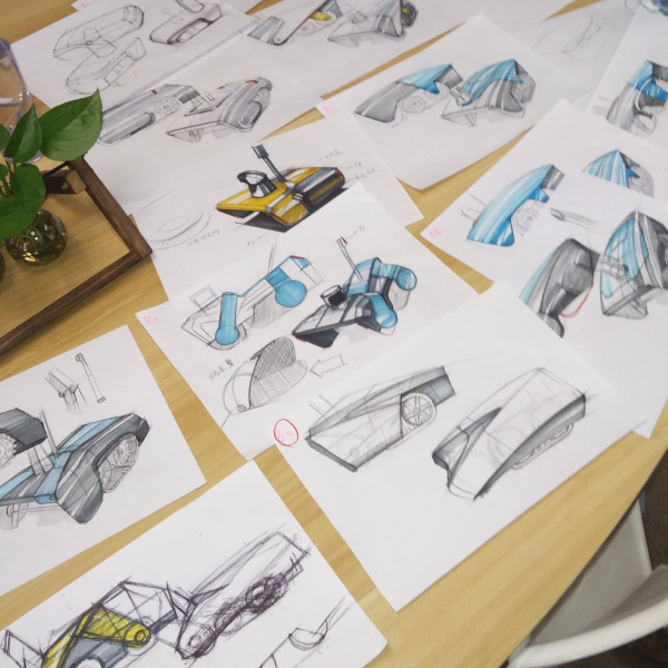 Sketches of futuristic vehicles in blue, gray, and yellow on white paper scattered across a wooden table, with a green plant in the corner.