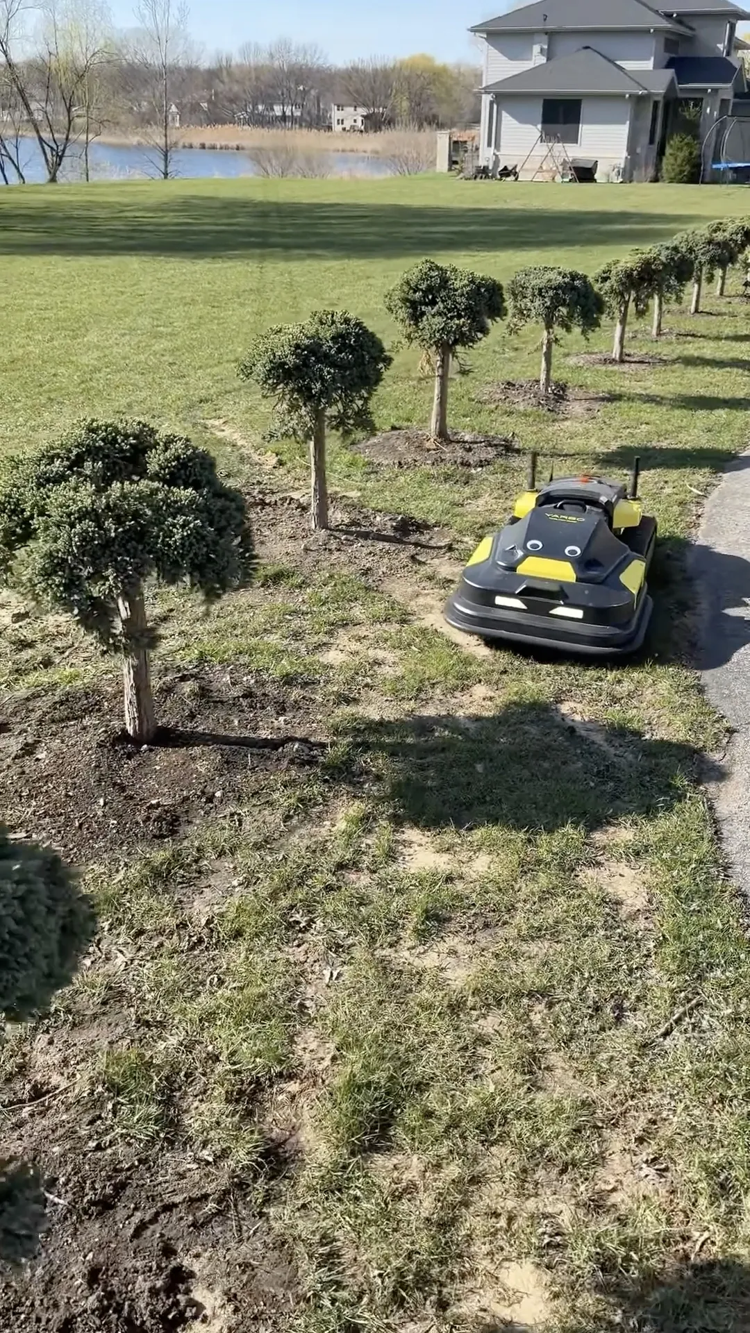 Black and yellow robotic lawn mower on grass near trimmed trees, with house and lake in background.