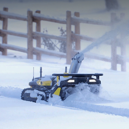 Yellow and black snow blower clearing snow in winter, wooden fence in background, snow-covered ground.