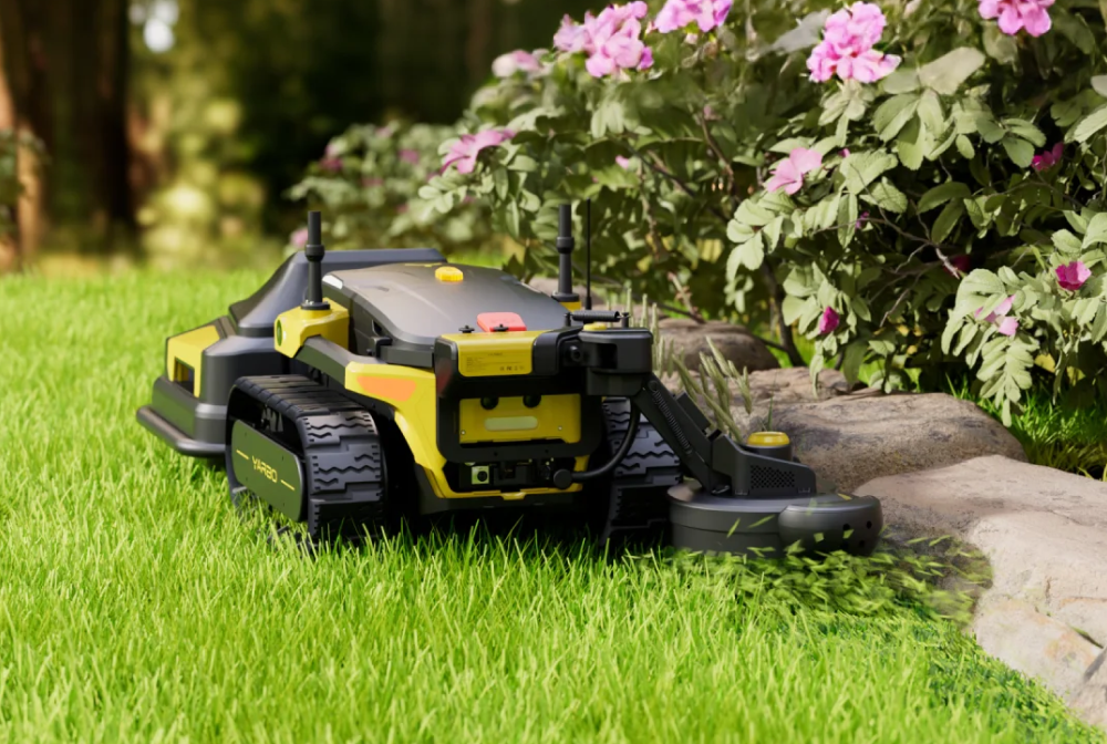 Yellow and black robotic lawn mower on green grass near pink flowers and rocks, cutting weeds.