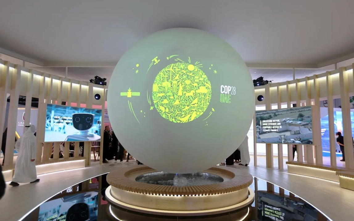 Large illuminated globe with green Earth design and "COP28 UAE" text, displayed in a modern exhibition space with screens and visitors.
