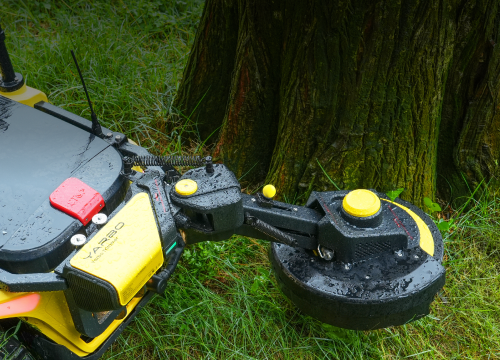 Yellow and black robotic lawn mower with circular cutting blade near tree trunk in grassy area.