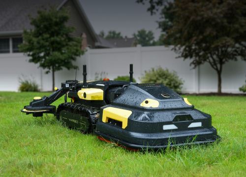 Black and yellow robotic lawn mower on green grass, with white fence and trees in background.