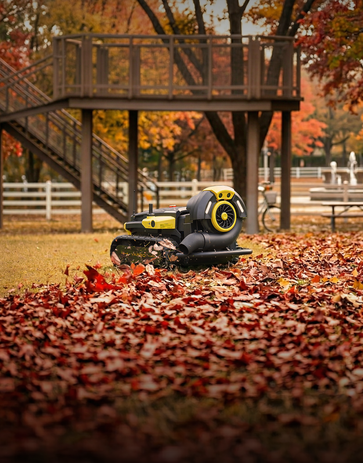 Black and yellow robotic leaf blower on grass covered in red autumn leaves, with wooden deck and trees in background.