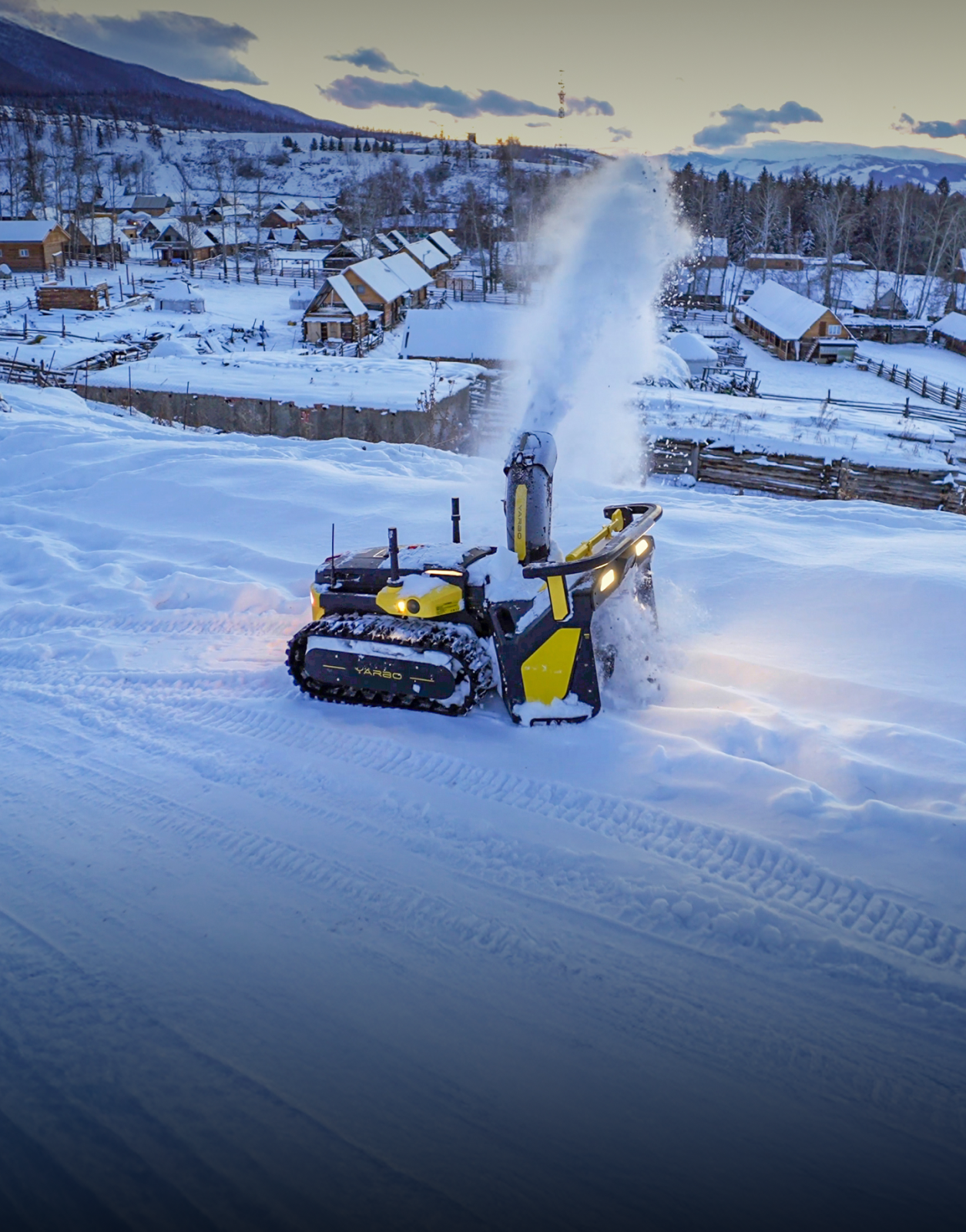 Yellow and black snow-clearing robot blows snow in snowy village at dusk, tracks visible, wooden cabins in background.