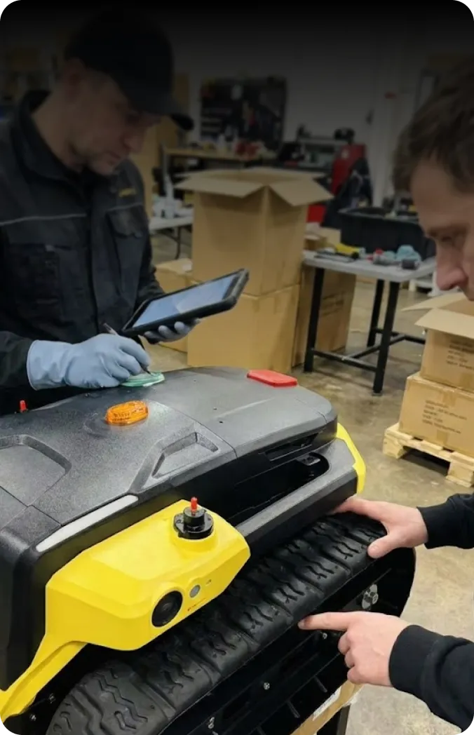 Two technicians inspect a yellow and black robotic lawn mower in a workshop, one using a tablet, the other pointing at its track.