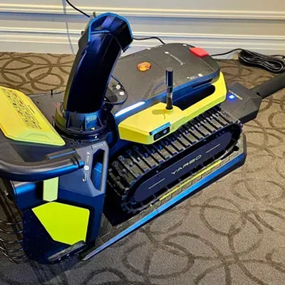 Blue and neon yellow robotic vacuum with tank treads on patterned carpet, power cord visible, illuminated blue light.