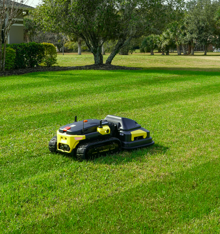 Yellow and black robotic lawn mower on green grass, trees and house in background, sunny day.