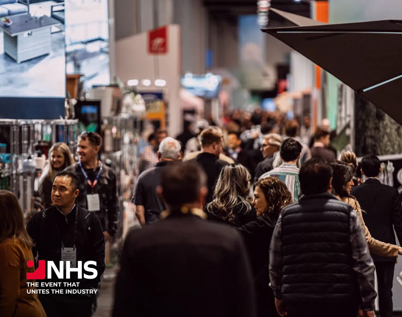 Crowd at NHS trade show, attendees walking between booths, logo visible, warm indoor lighting.