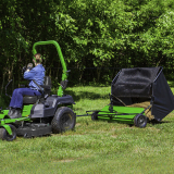 Person operating green riding lawn mower with attached black grass catcher on grassy lawn, trees in background.