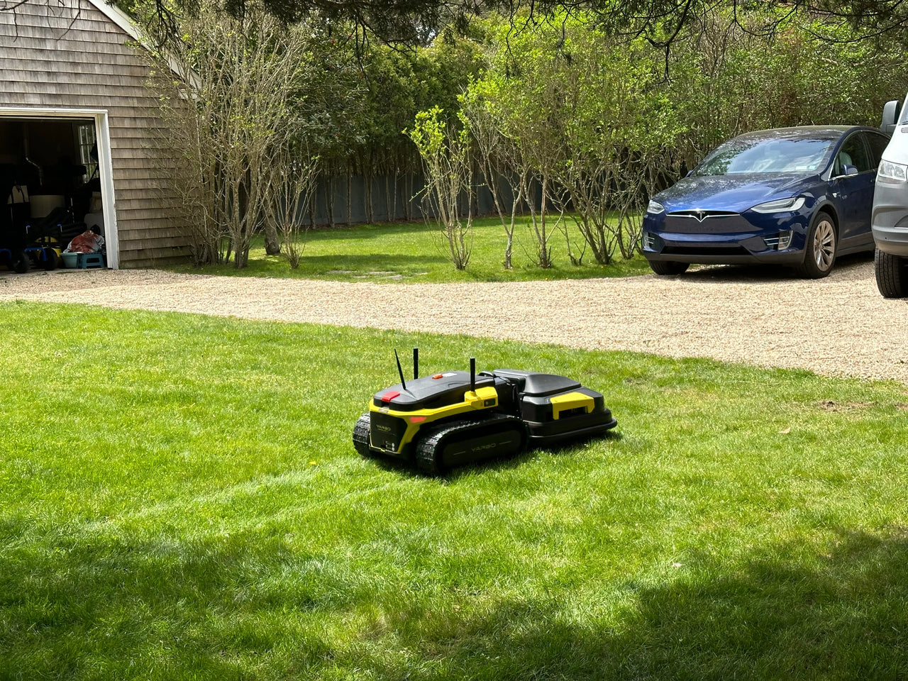 Robotic lawn mower on vibrant green grass in sunny suburban yard, near Tesla and garage.