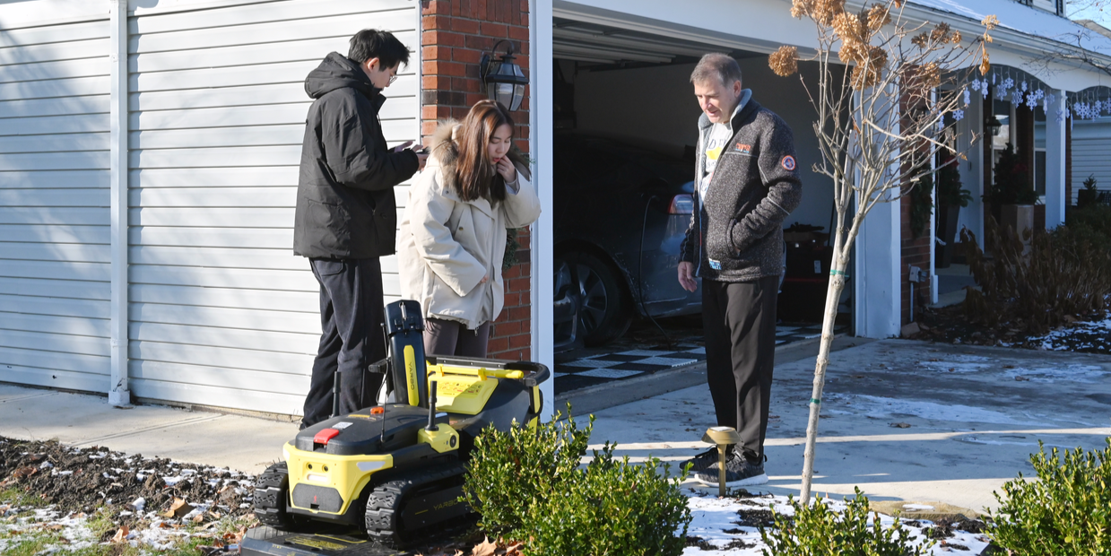 Mike Motsis stands by his home with two young people, smiling near a yellow robotic lawn mower on a snowy day.