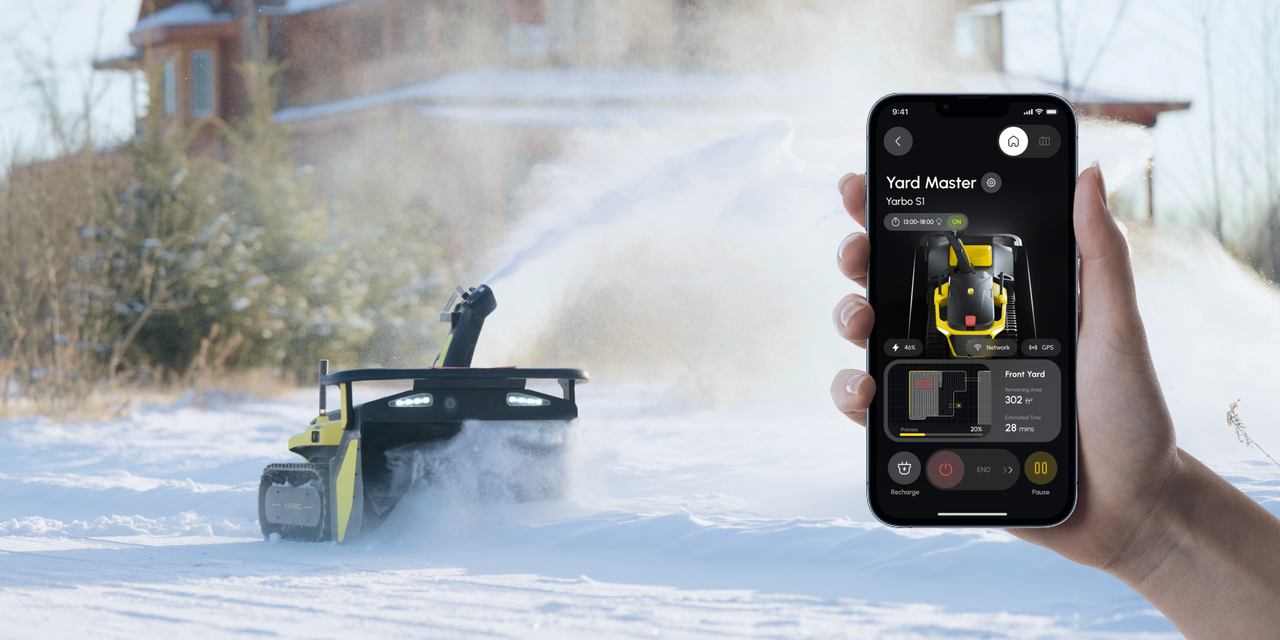 Hand holds phone controlling yellow remote snow blower clearing driveway in snowy yard.