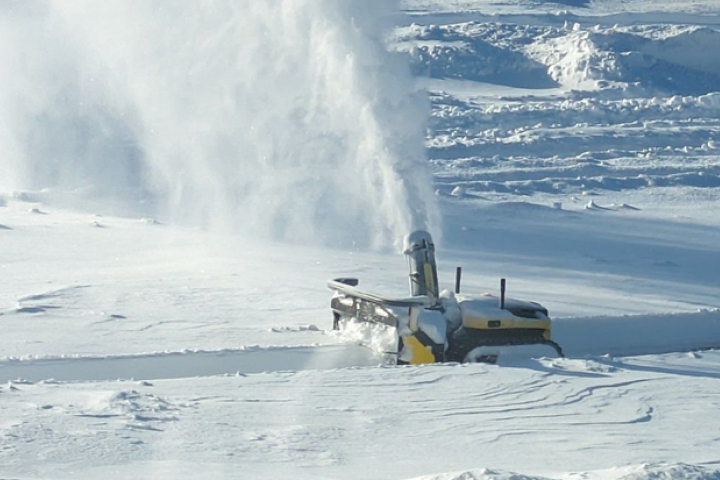 Yellow snow blower clears deep snow in bright winter sun, sending plume of snow high into air.