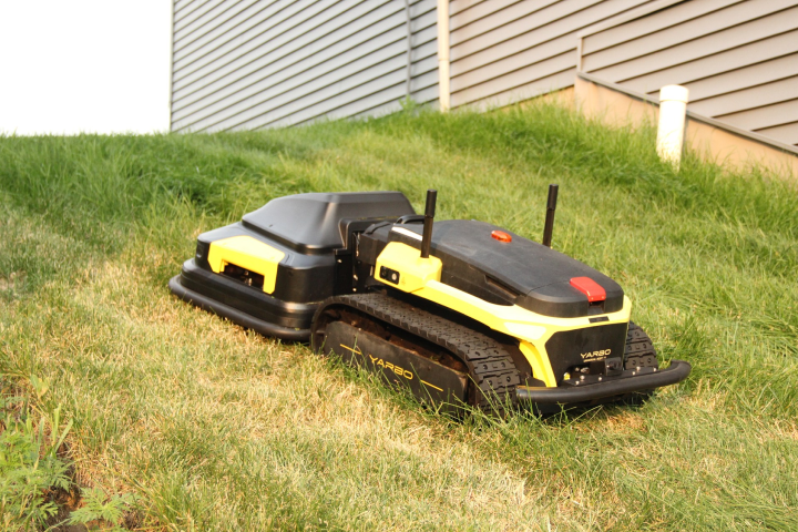 Yellow and black Yarbo robot lawn mower on grass near house, sleek design with tracks and antennas, ready for automated mowing.