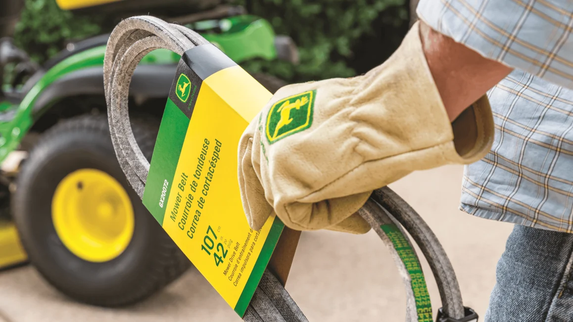 Gloved hand holding John Deere mower belt package, green and yellow, with lawn mower in background.