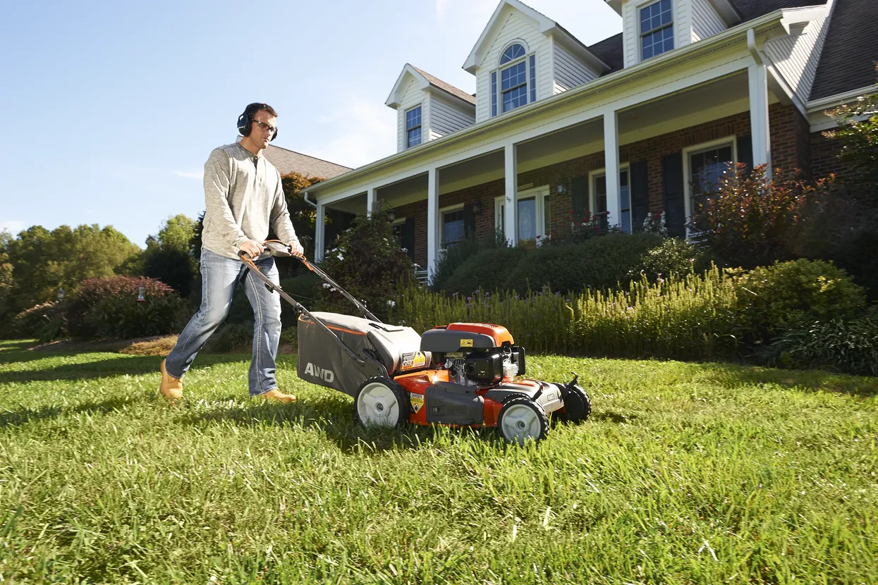 Man pushing manual lawn mower on sunny day, trimming lush green grass in front of suburban home.