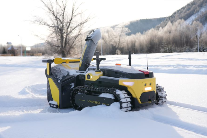 Yellow and black YARBO robotic snow blower clearing snow on a sunny winter day, with tracks and anti-clog chute visible.