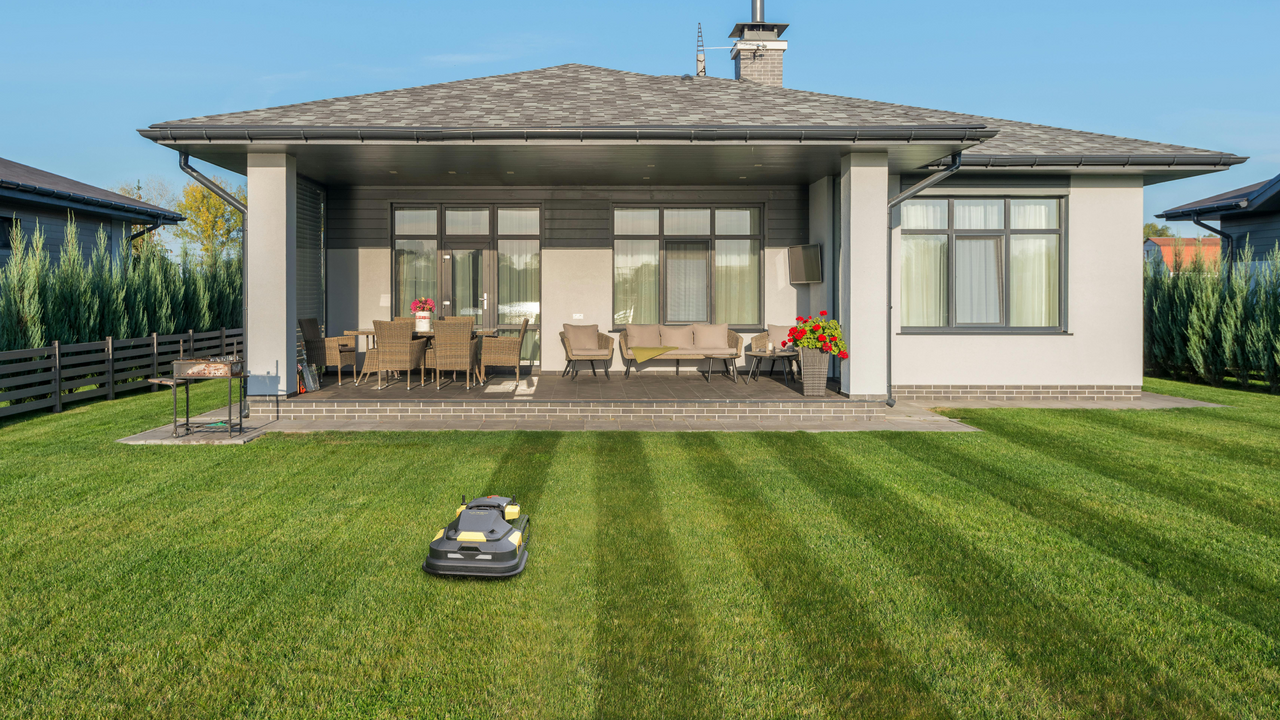 Robotic lawn mower glides across lush green lawn in front of modern home with patio and outdoor furniture.