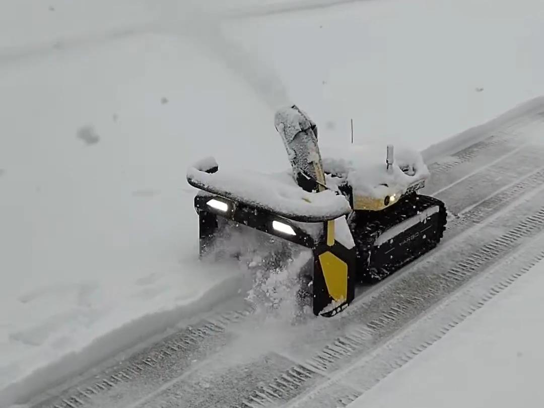 Robotic snow blower clears driveway in heavy snow, bright lights glowing, tracks cutting through deep white snow.